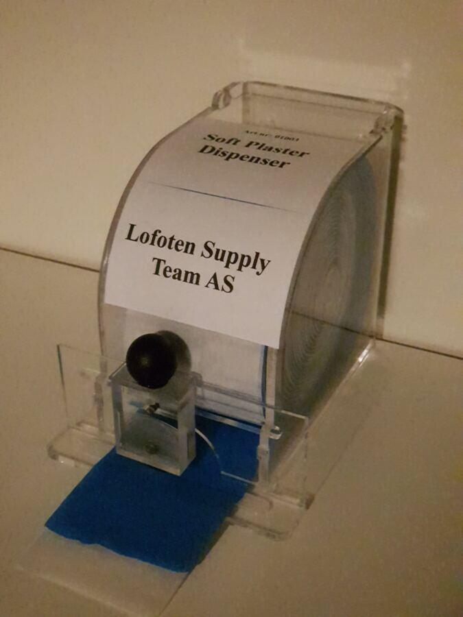 Dispenser i akryl for bandasjeplaster, merket Lofoten Supply Team, med bl&aring;tt plaster og sirkelkniv